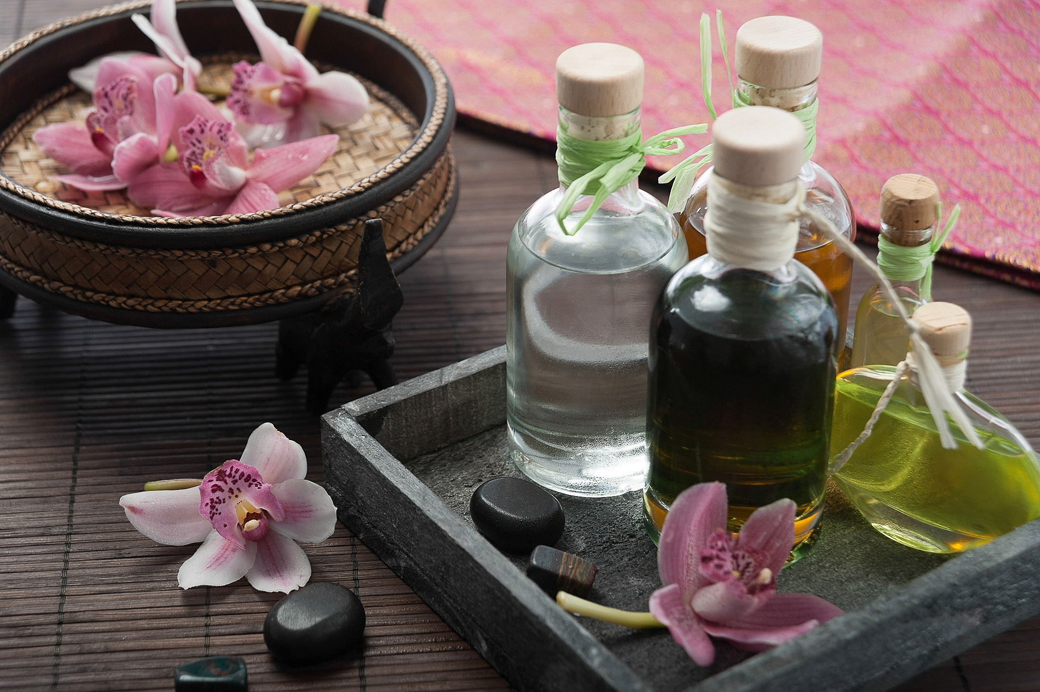 Orchid flower with white and pink petals resting on a bamboo mat, surrounded by soft natural light. The background includes a wooden bowl and a stone tray, creating a calm and serene atmosphere. No visible text. The overall mood is peaceful and elegant.